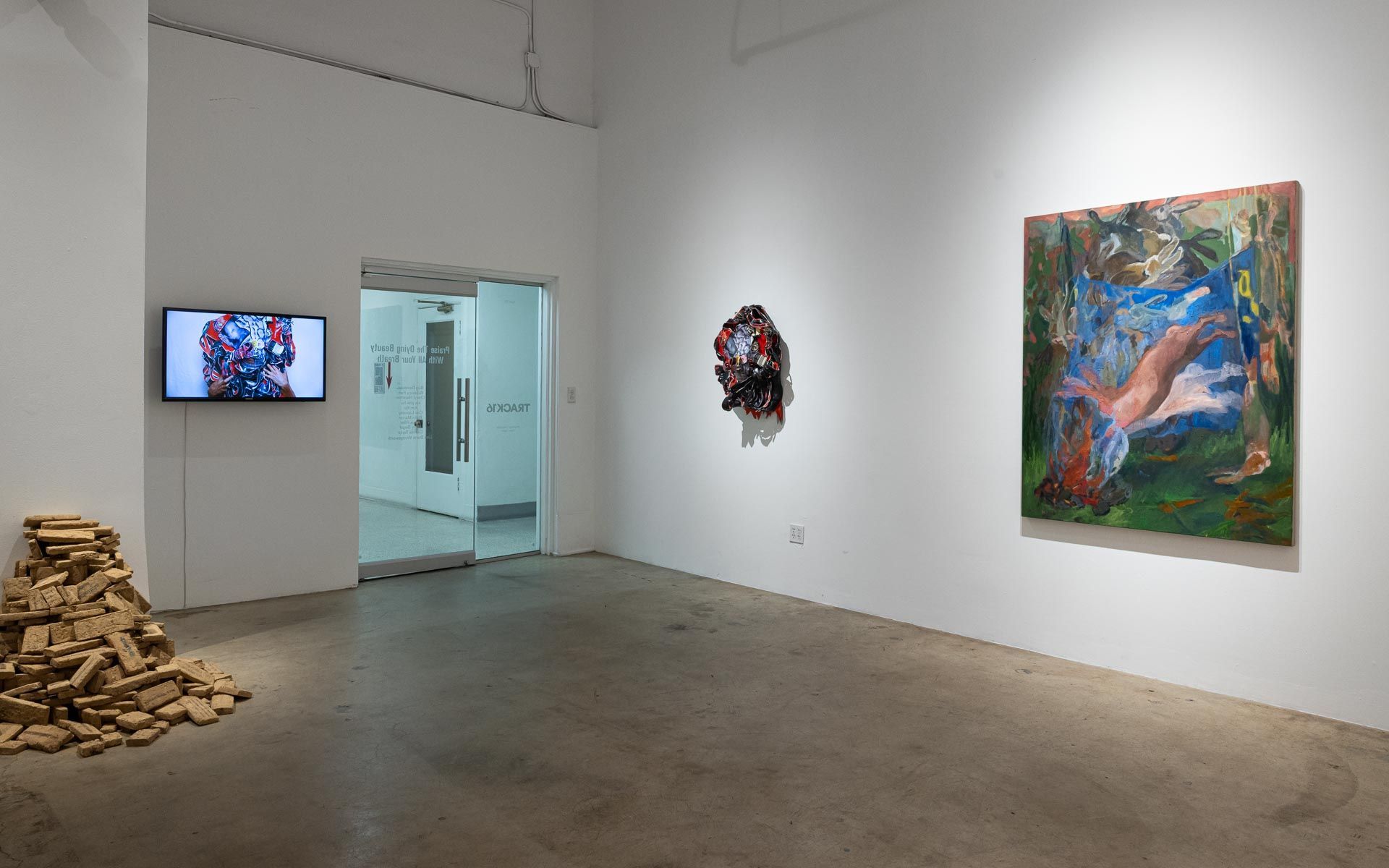 Art gallery with a TV on the wall, stacked wood at left, and colorful paintings on white walls. Praise the Dying Beauty art exhibition at Track 16 Downtown Los Angeles gallery. Curated by Galia Linn featuring sculpture, paintings and drawings. 