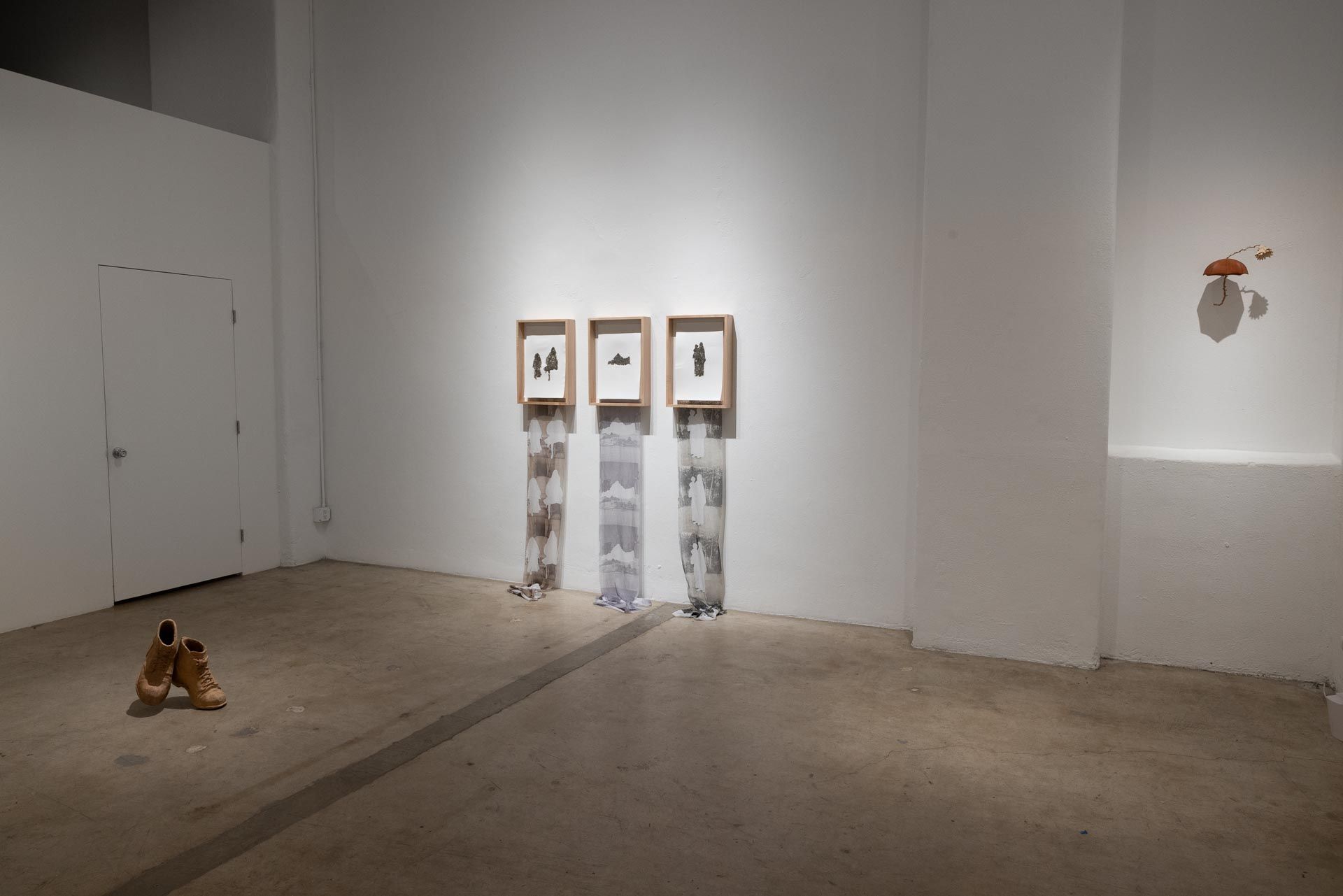 Three framed drawings of figures with long printed fabric strips hanging below, a pair of tan boots on the floor and a white door at left. Installation view, Valence: Azadeh Shladovsky & Daniel Wheeler, Track 16.