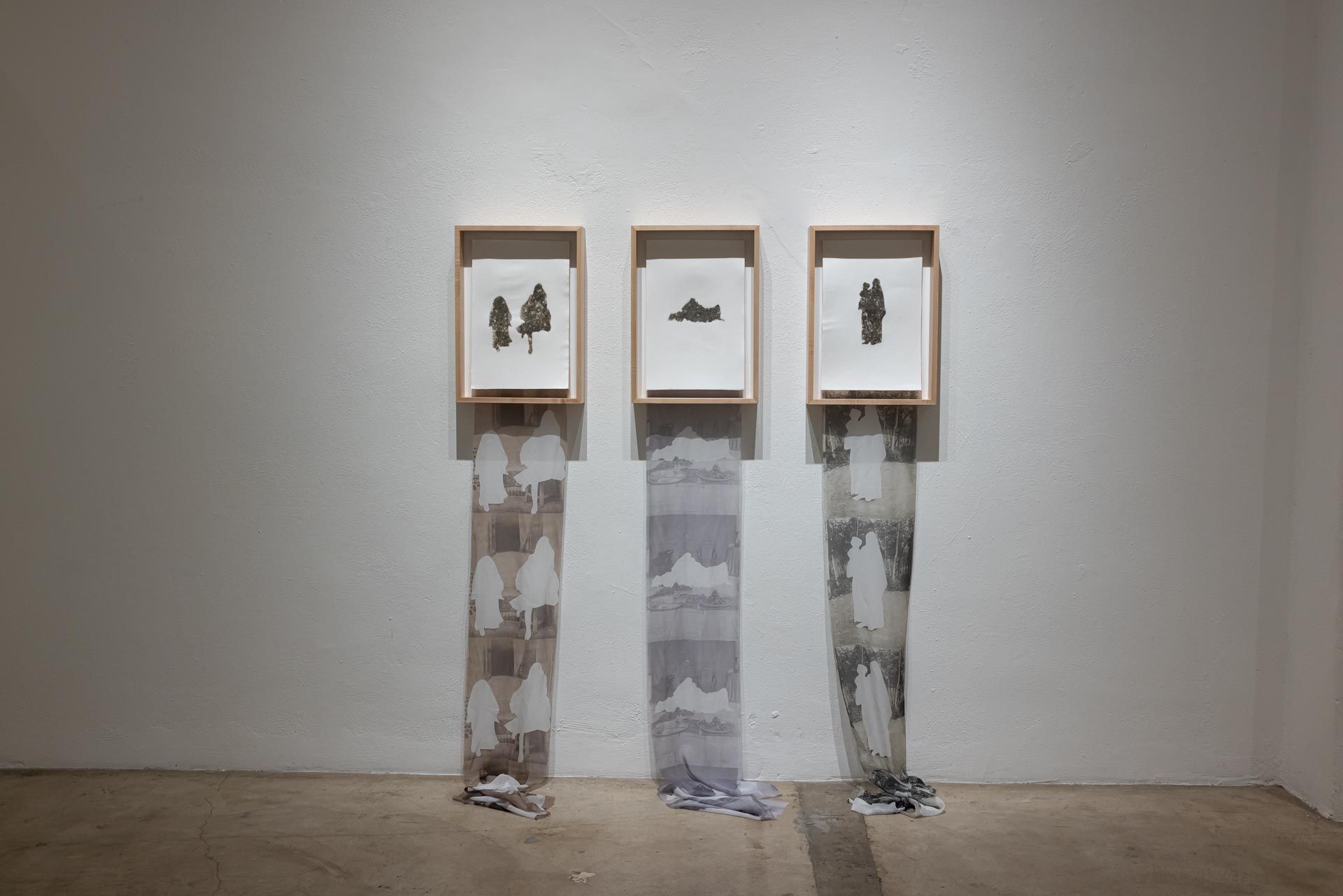 Close view of three wood-framed drawings of dark silhouettes, each with a long gray-and-white printed fabric strip hanging to the floor. Installation view, Valence: Azadeh Shladovsky & Daniel Wheeler, Track 16.