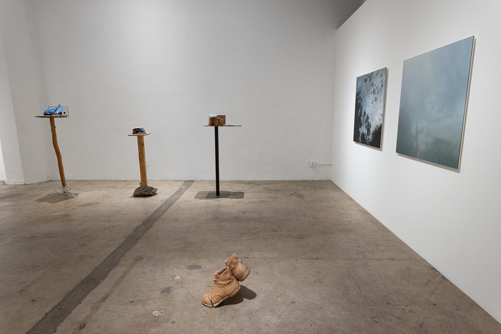Three wood and metal stands with small sculptures at left, two blue-gray paintings on the right wall and a sculpted boot on the floor. Installation view, Valence: Azadeh Shladovsky & Daniel Wheeler, Track 16.