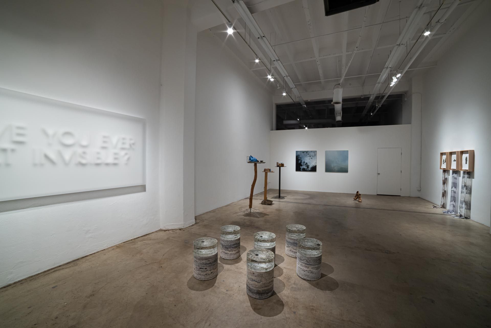 Wide view of a long gallery: oversized white relief text, stacked paper discs, sculpture stands, two paintings and framed fabric works at right. Installation view, Valence: Azadeh Shladovsky & Daniel Wheeler, Track 16.