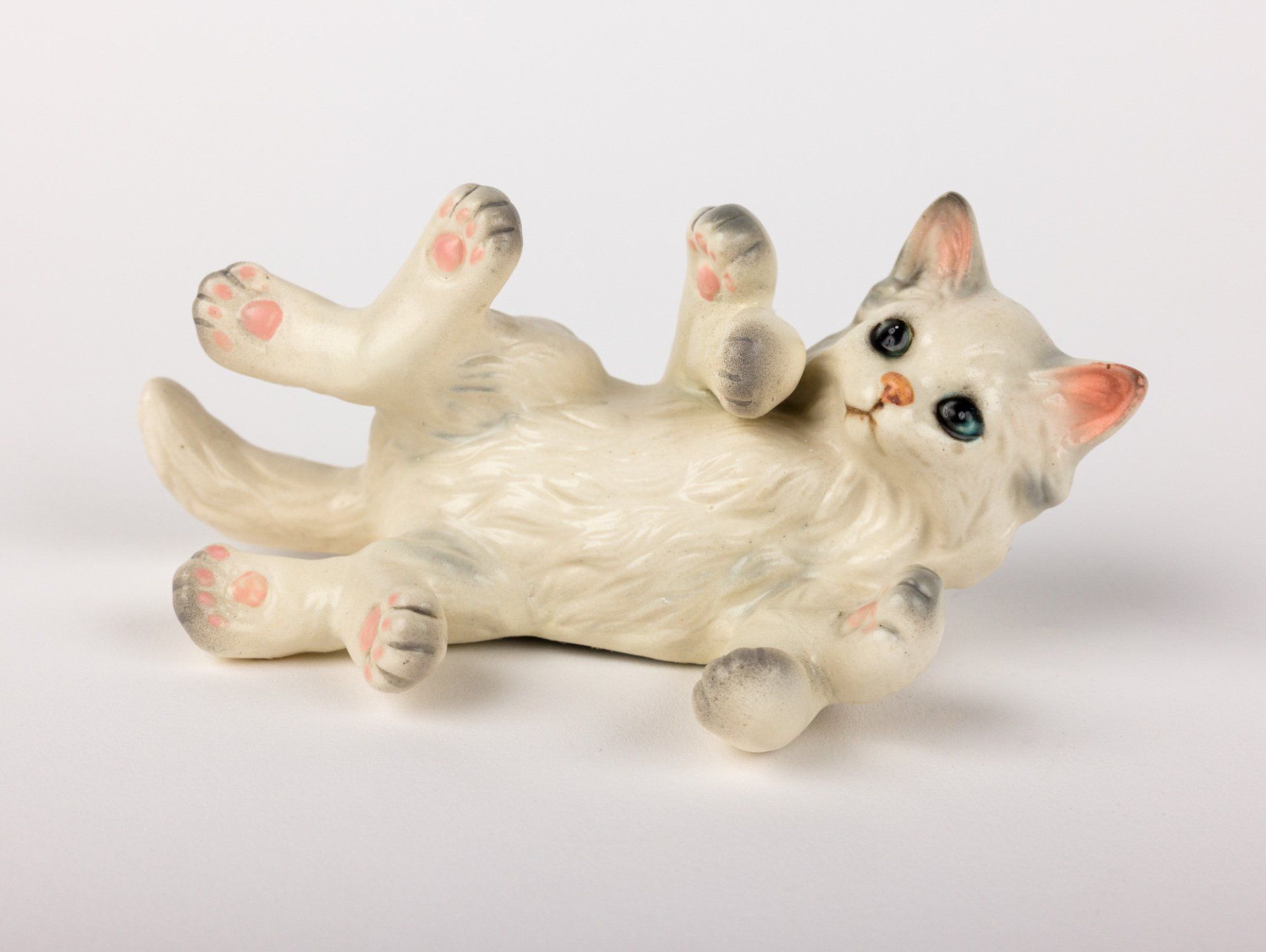 A white cat figurine is laying on its back on a white surface.