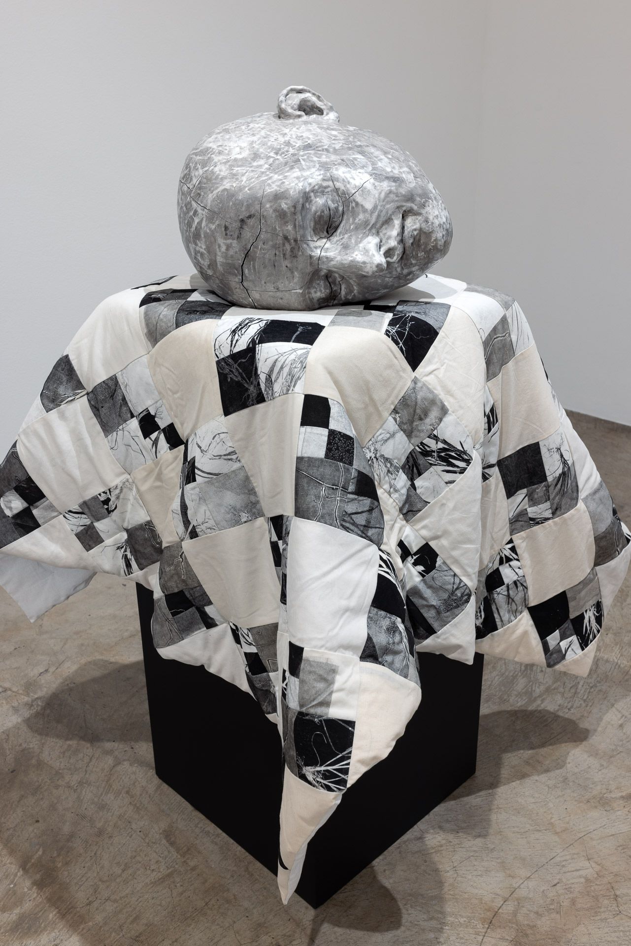 Gray textured sculpture on a black pedestal draped with a black-and-white patchwork cloth in a gallery setting