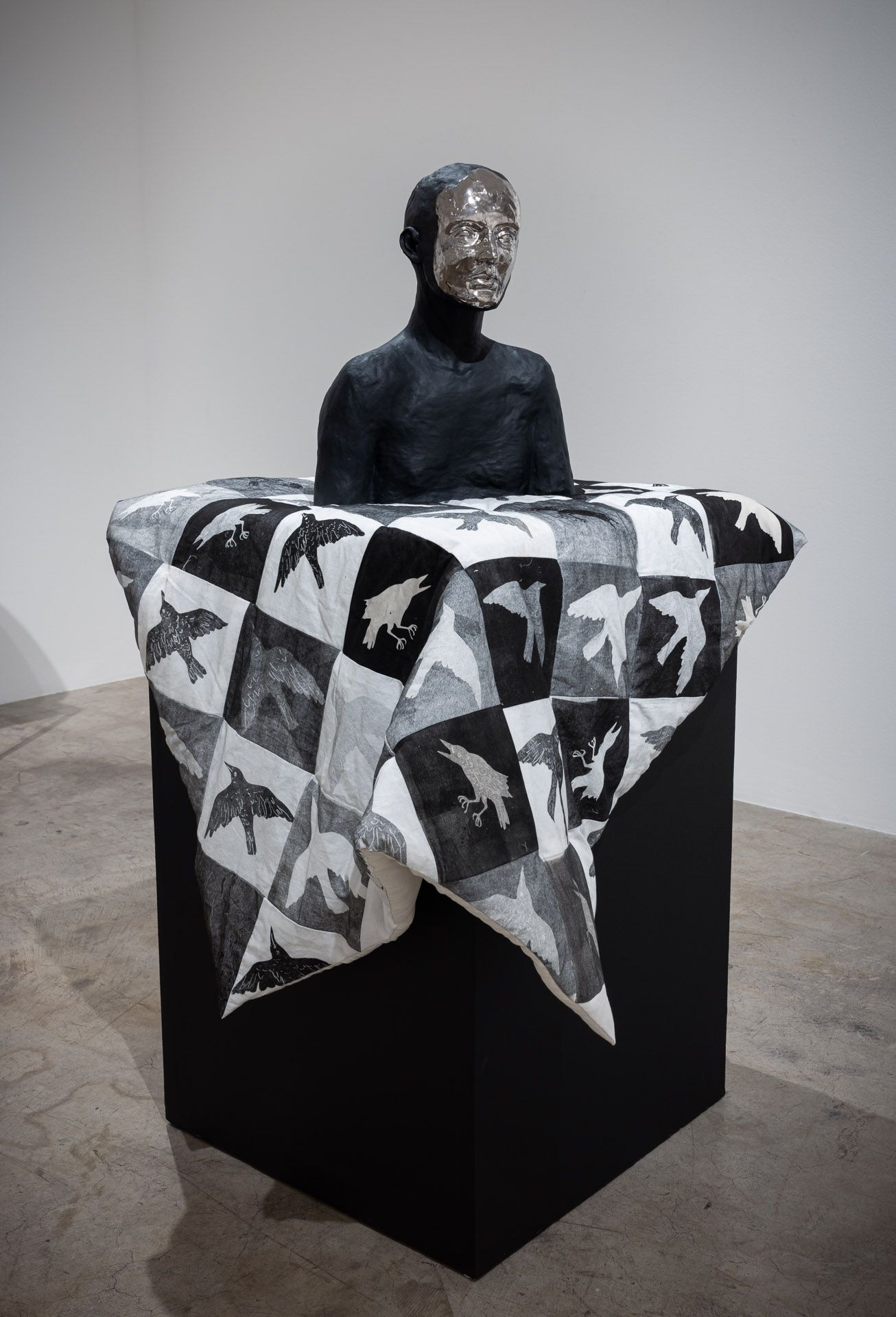 Abstract sculpture on a black pedestal, draped with a black-and-white geometric cloth in a gallery.