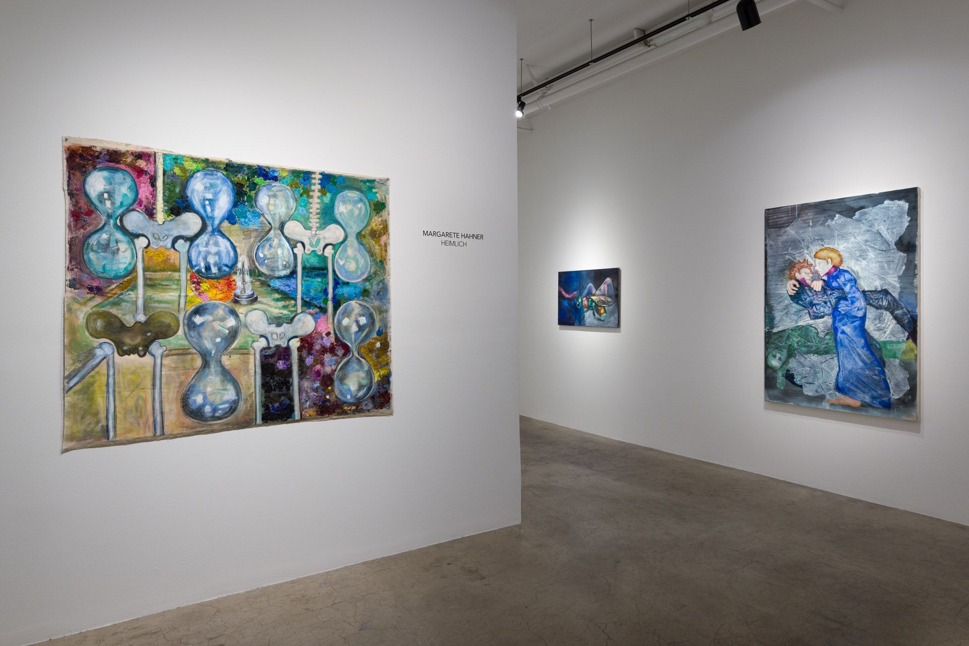 Large painting of blue hourglass shapes beside title wall text, a small painting and a painting of a figure in a blue robe on the wall beyond. Installation view, Margarete Hahner: Heimlich, Track 16.