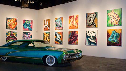A green car is parked in a room with paintings on the wall.