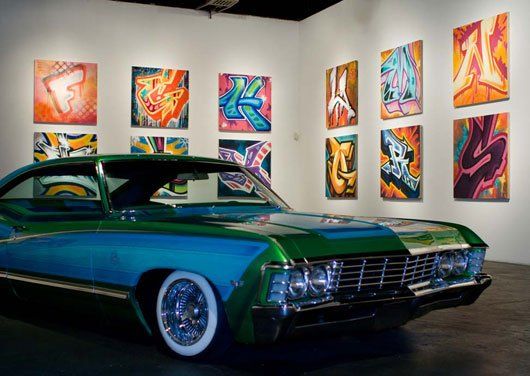 A blue and green car is parked in front of a wall of paintings.