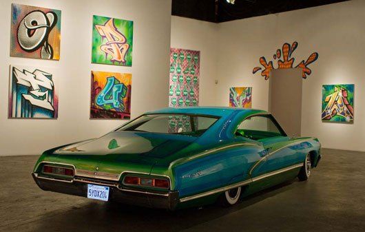 A green and blue car is parked in a room with paintings on the wall.