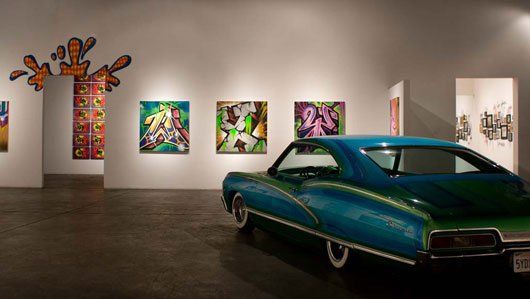 A blue and green car is parked in a room with paintings on the wall.