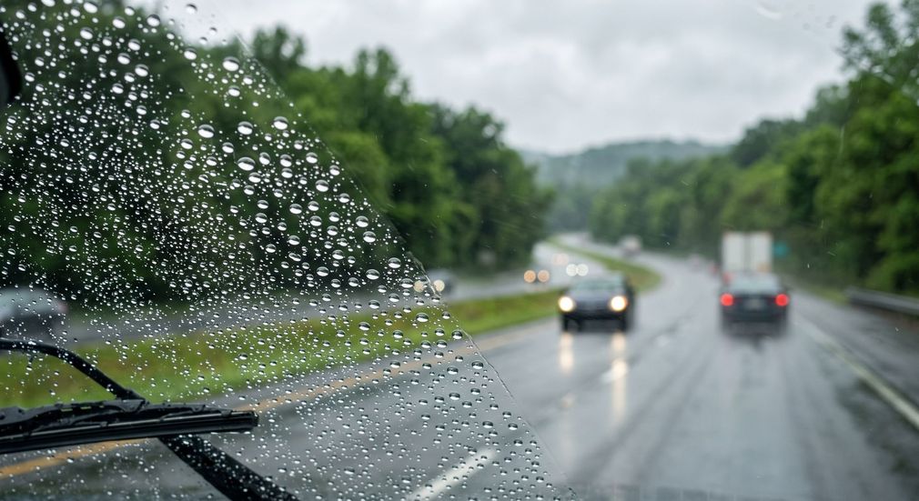 Rainy highway seen through windshield with wipers, blurred cars ahead, and wet road surrounded by trees