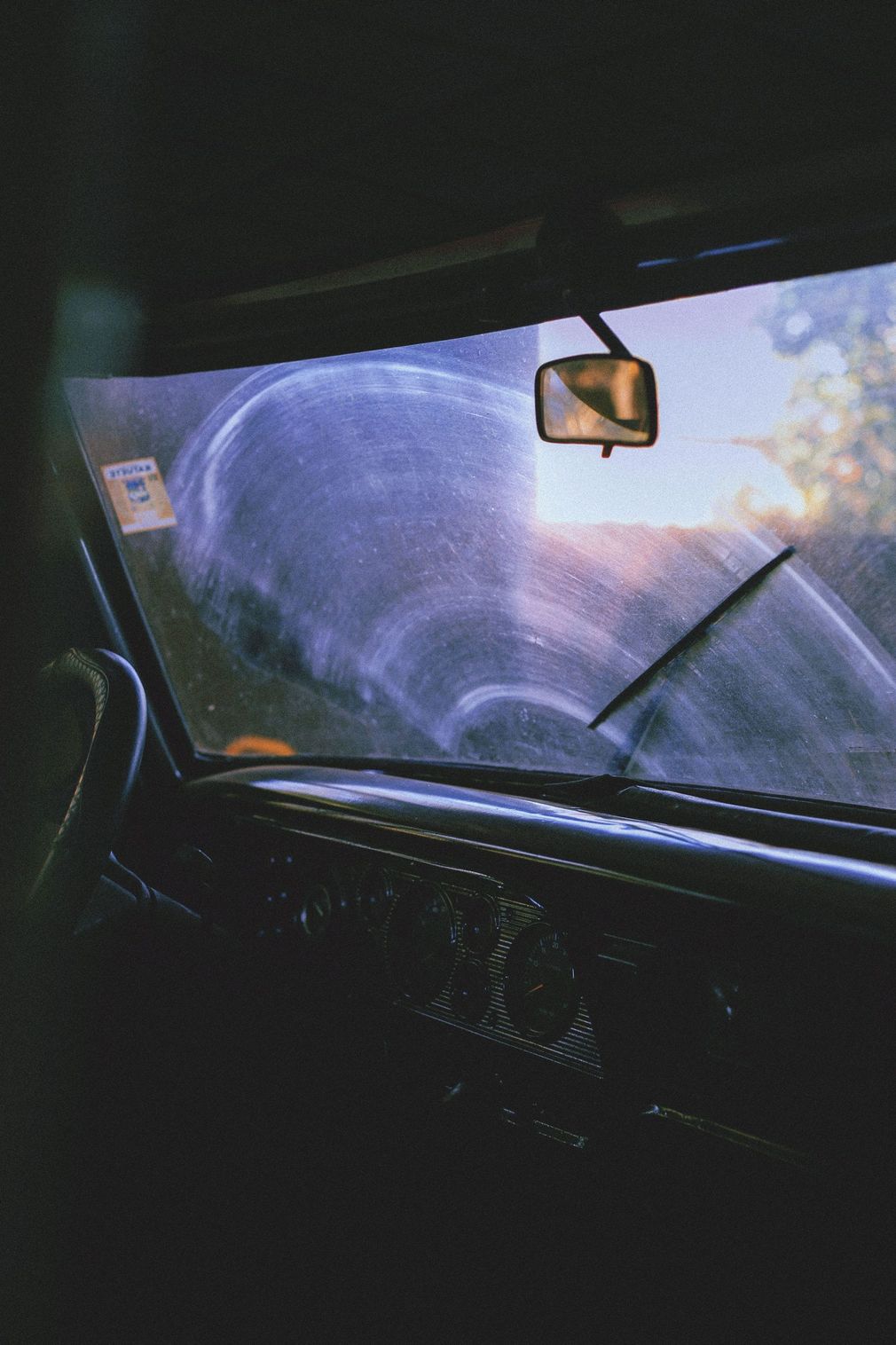 Windshield being washed from inside a dark car, with sunlight and a rearview mirror visible.