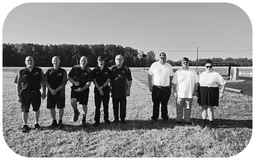Group of people posing on a grassy field, with trees and buildings in the background. | Dewitt's Automotive Center
