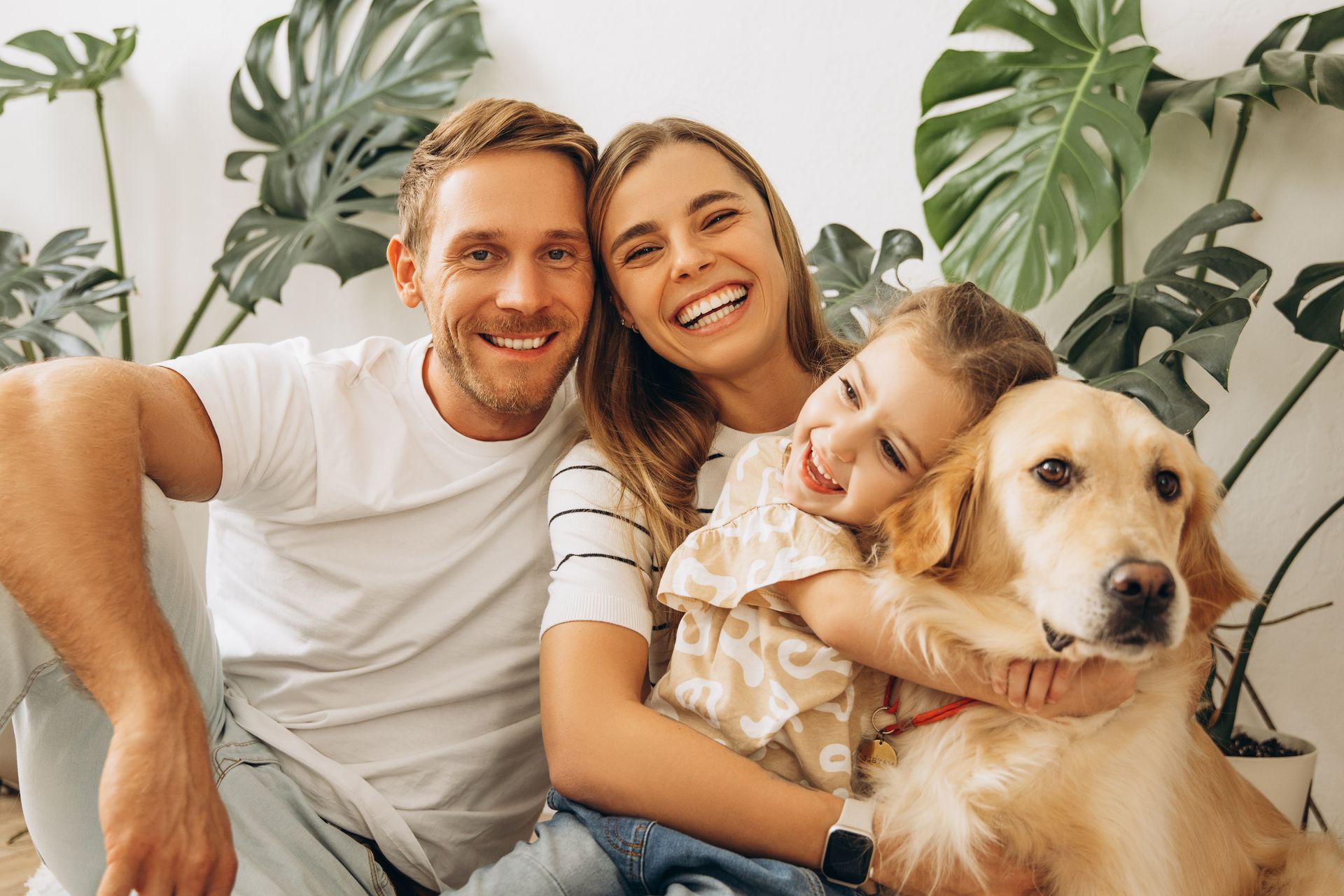 Family of three and golden retriever smiling in front of monstera plants