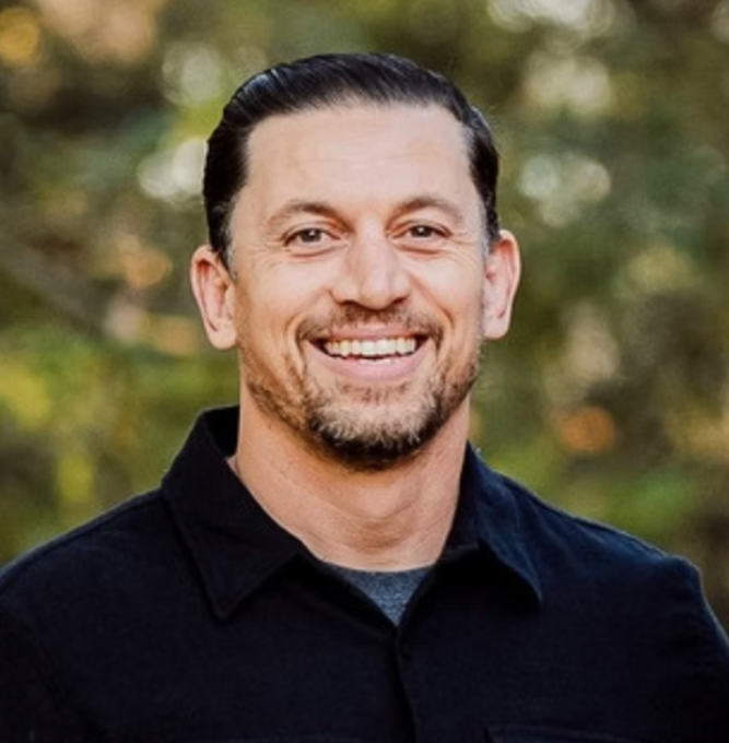 Smiling man in a black shirt outdoors with a blurred green background