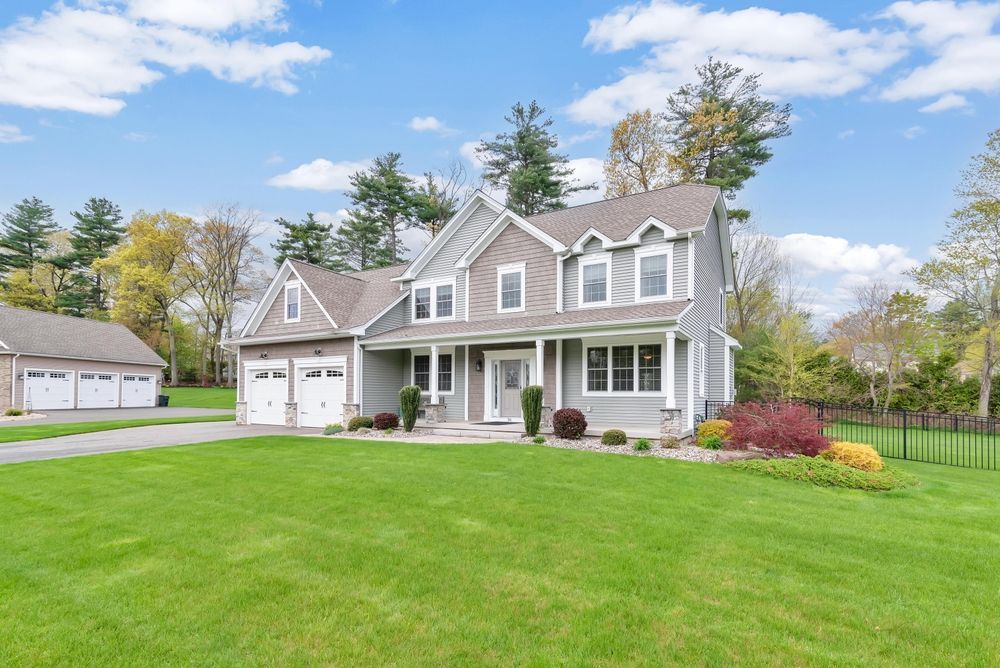Suburban two-story gray house with white trim, attached garage, and a large green lawn under a blue sky