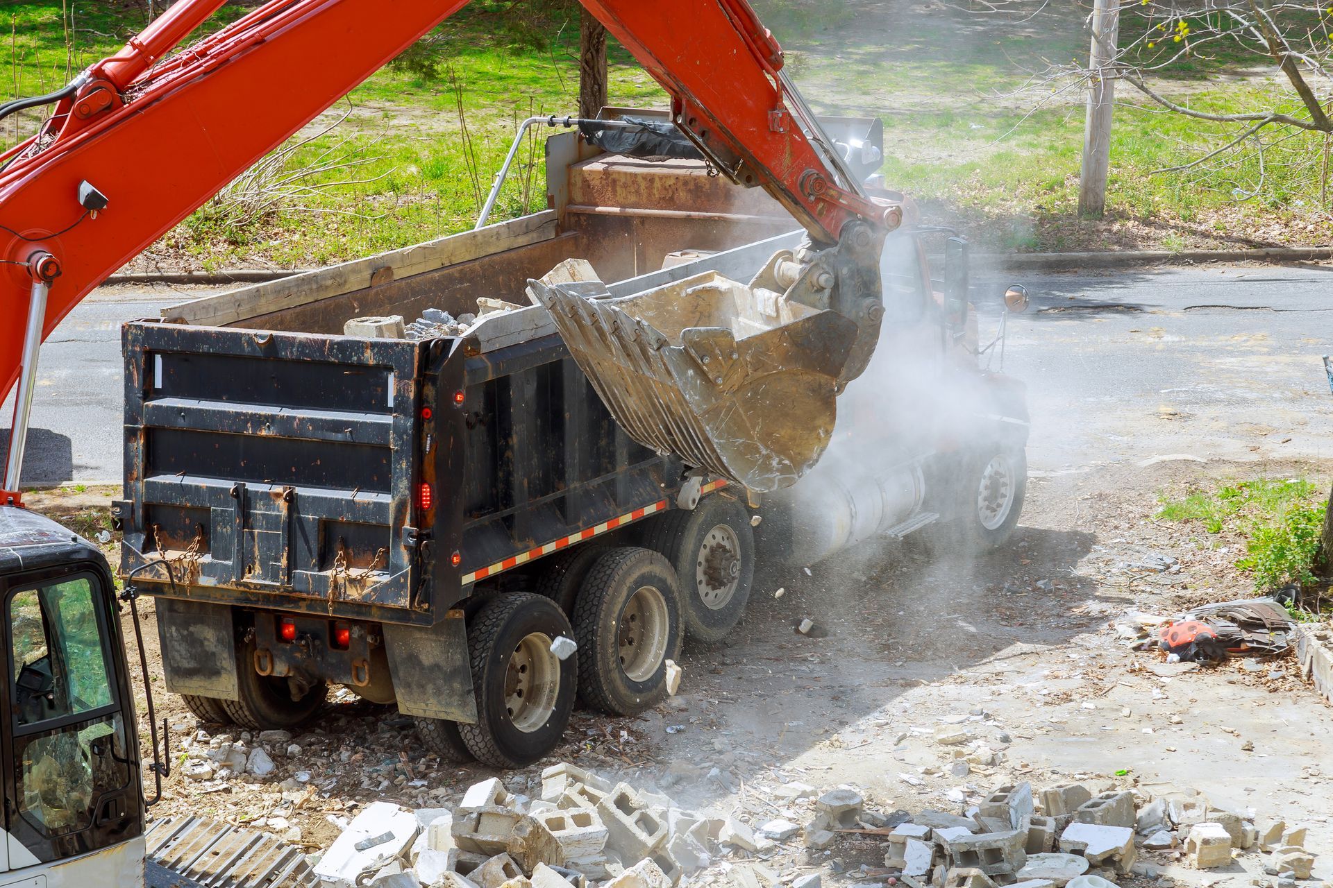 Orange excavator loading debris into a black dump truck outdoors, creating dust.