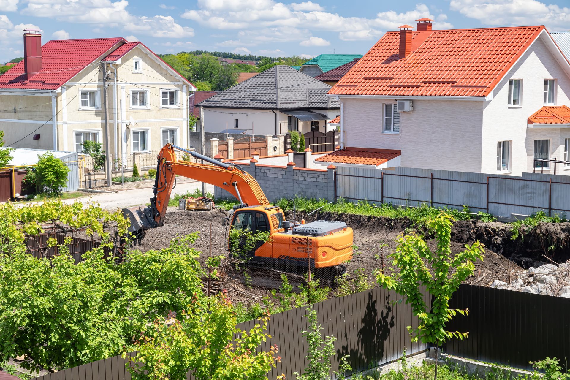 Orange excavator digging in a residential area, houses with red and orange roofs in the background.