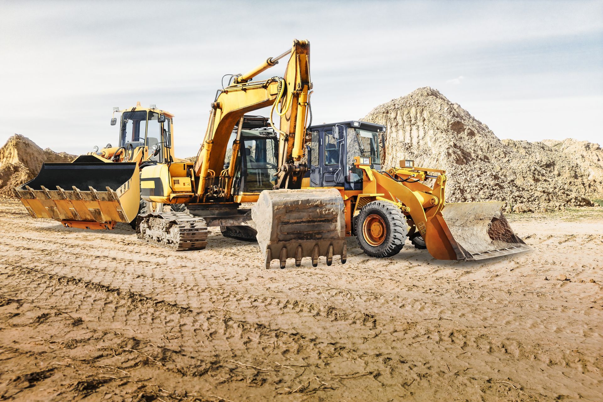 Three yellow construction vehicles on a dirt construction site near piles of dirt.
