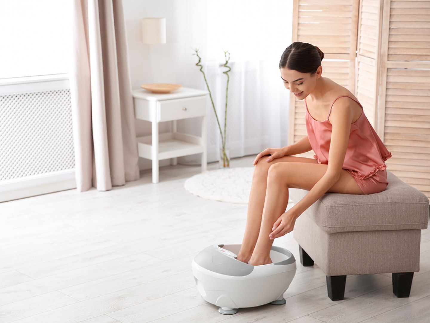 Woman using a white foot spa while seated on a beige ottoman in a bright room.