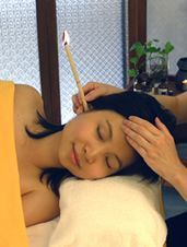 Person lying on a massage table with eyes closed while another hand holds a candle nearby