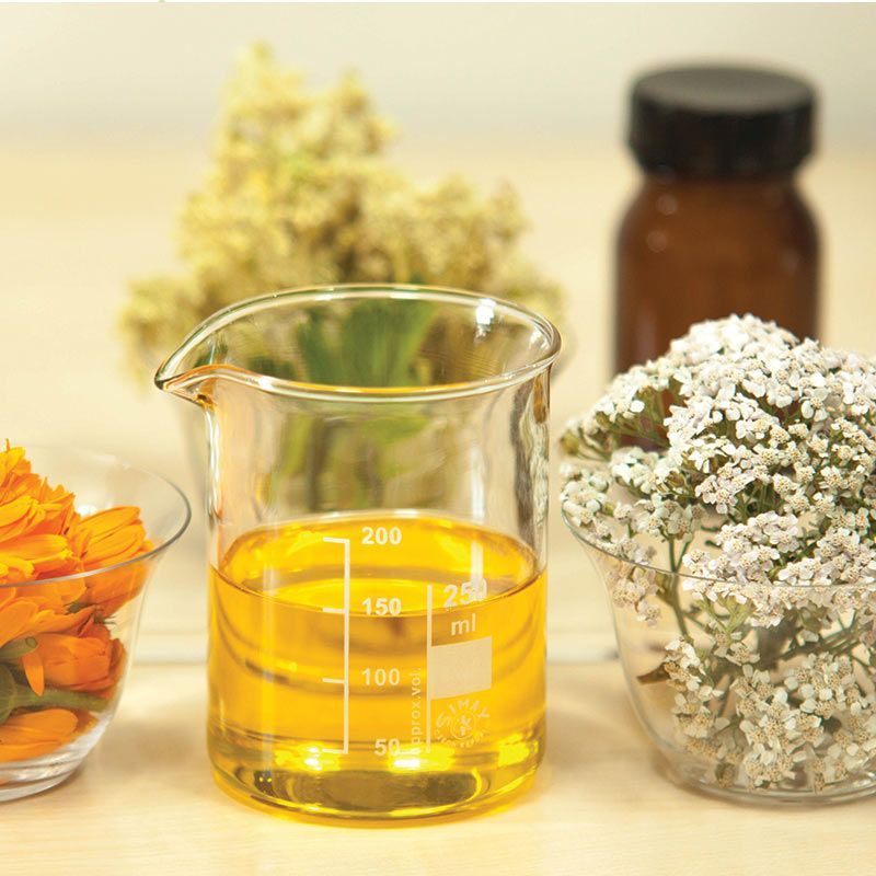 Clear beaker of yellow herbal oil with flowers and dried blossoms on a table