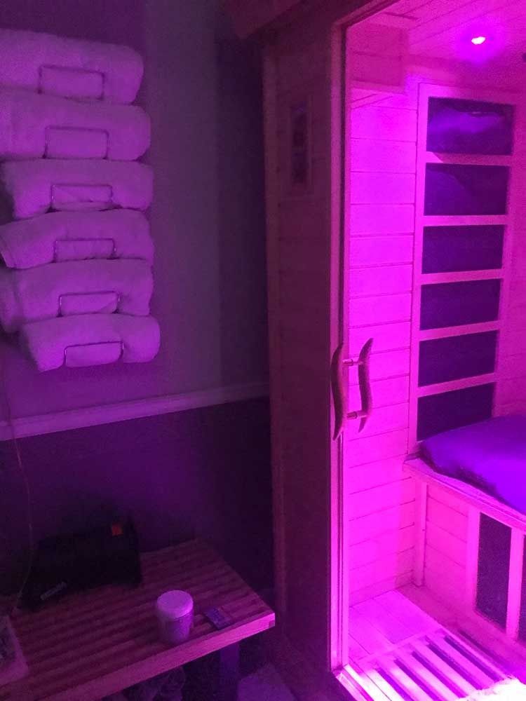 Purple-lit interior with stacked folded towels, a bench, and a doorway to a sauna-like room