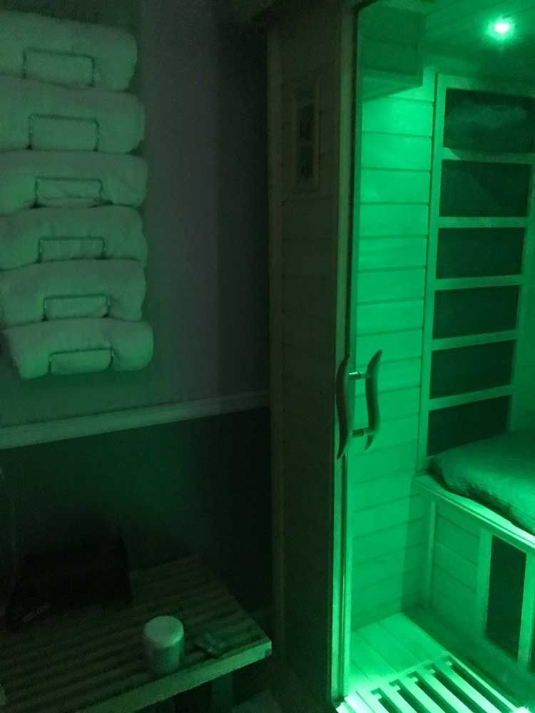 Green-lit sauna with benches, stacked towels, and a cup on a lower shelf.