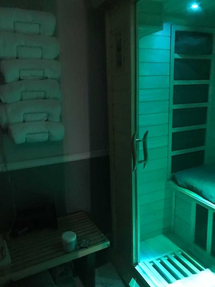 Green-lit indoor sauna with stacked towels and a glass door beside a bench and cupholder