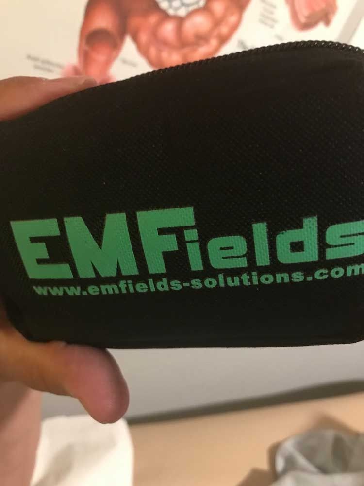 Black zip pouch with green “EMFields” logo held in a hand, against a blurred background.
