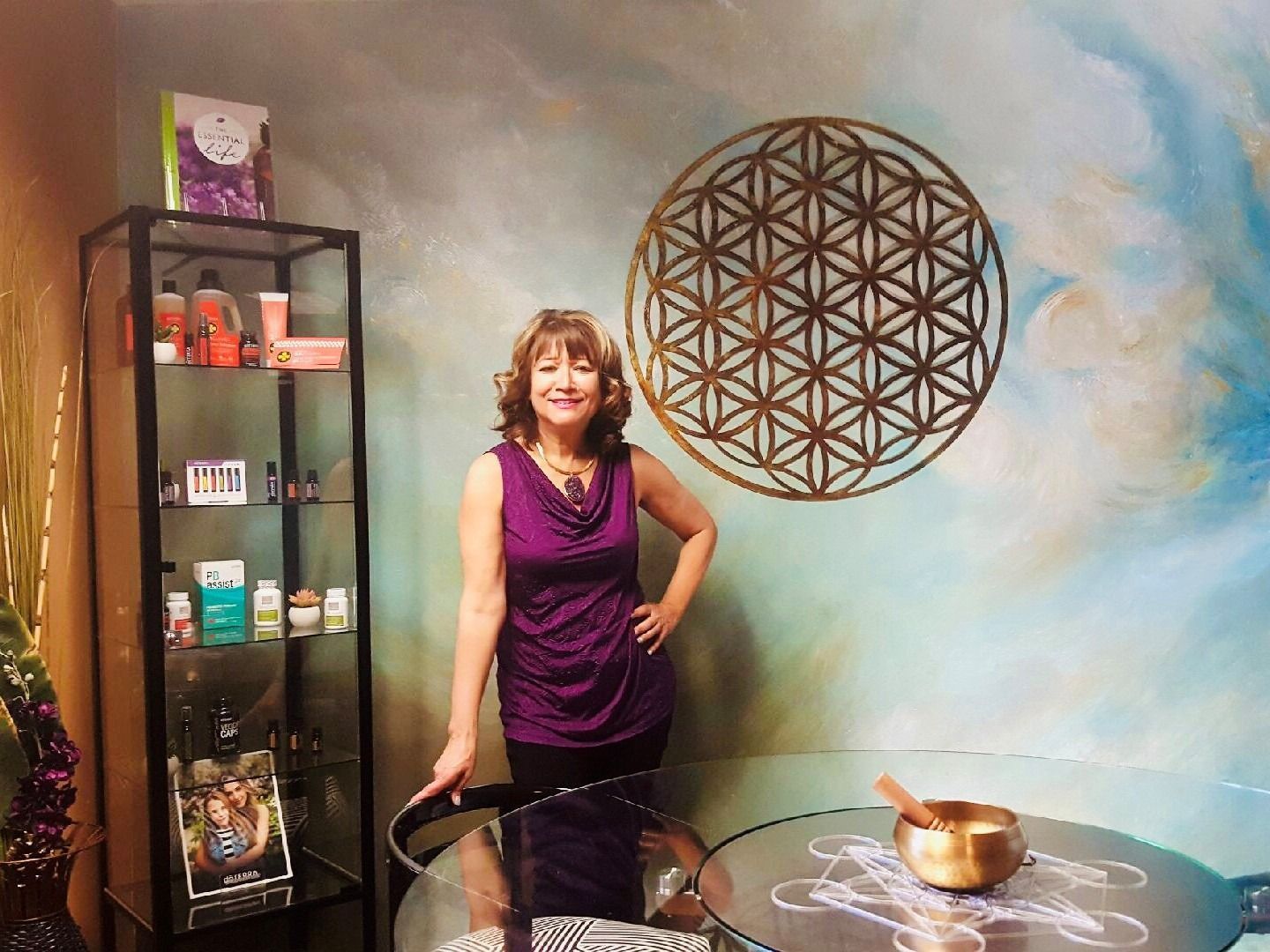Woman in a purple dress stands beside a glass table and display cabinet in a decorative room.