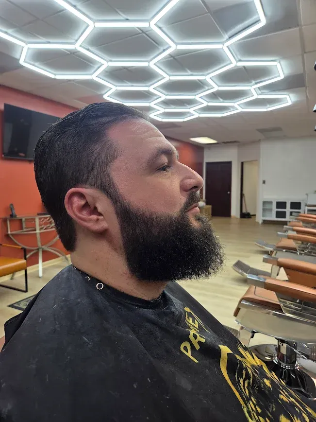 Side-by-side photos of a man with a fresh fade haircut, styled with a hard part and gel, wearing a patterned barber cape.