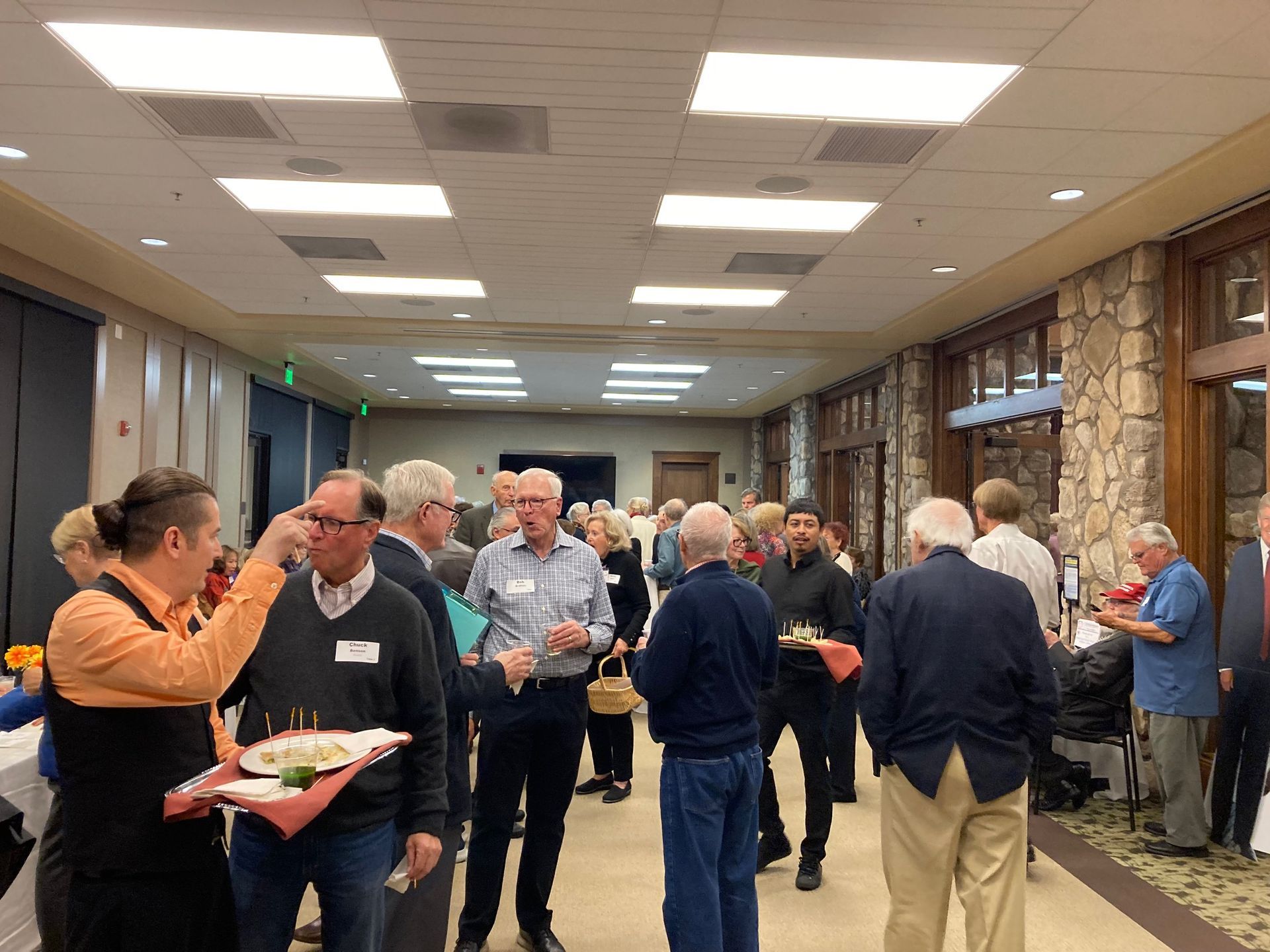 People mingling at a conference reception in a hotel hallway, holding drinks and chatting under bright ceiling lights