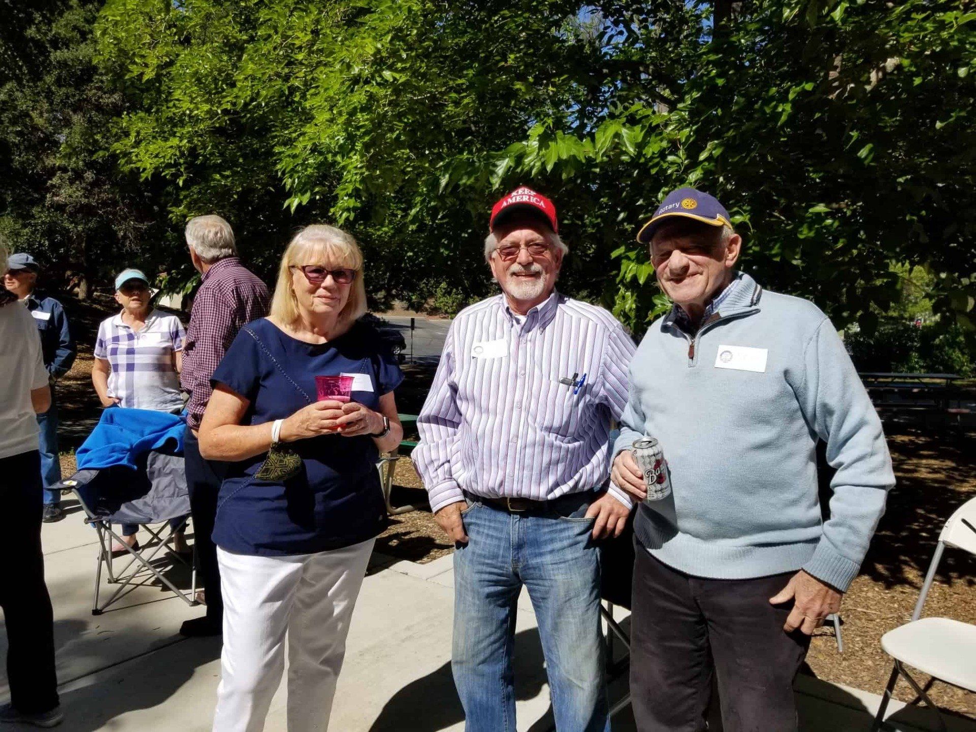 Three people standing outdoors, smiling and holding drinks at a sunny gathering