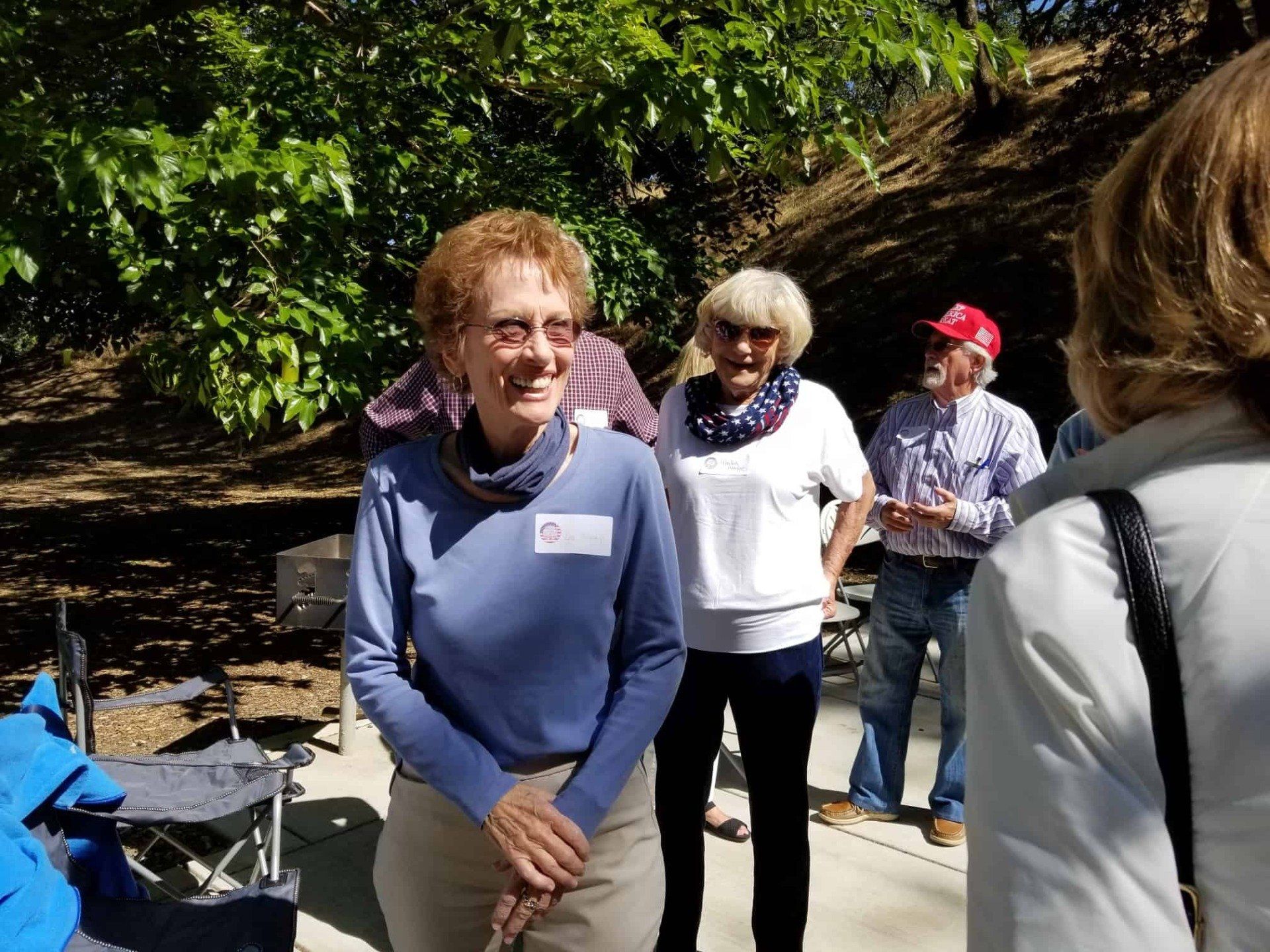 People standing outdoors in a sunny wooded area, with one smiling woman in a blue sweater in front