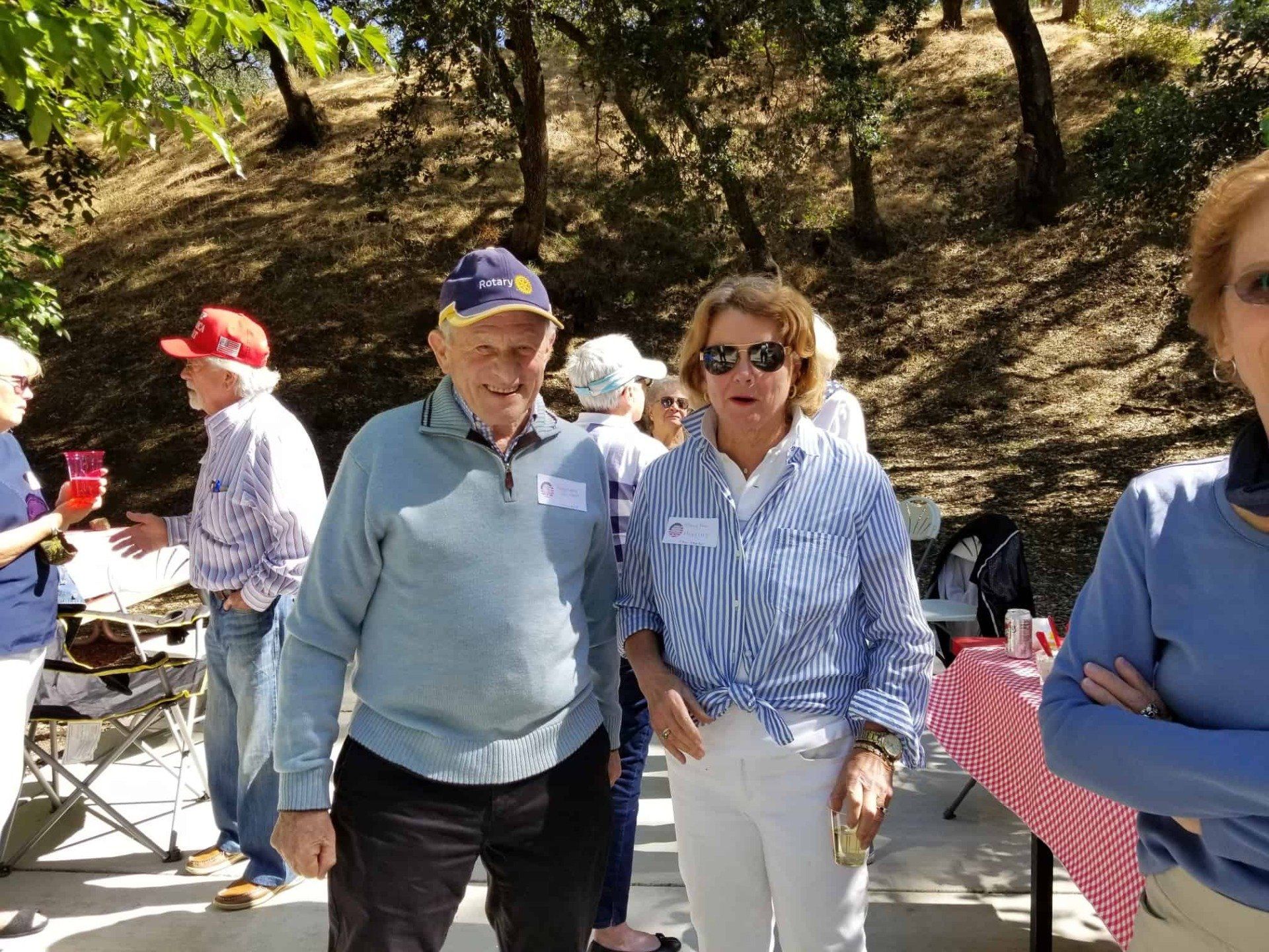 People standing outdoors at a picnic table under trees, with drinks and casual clothes.
