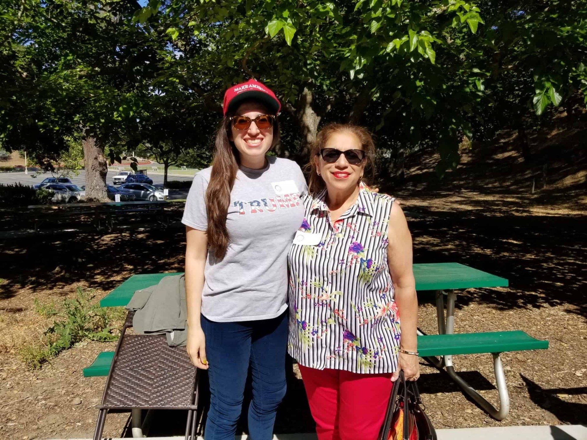 Two women smiling outdoors by a picnic table under trees, wearing sunglasses and casual clothes.