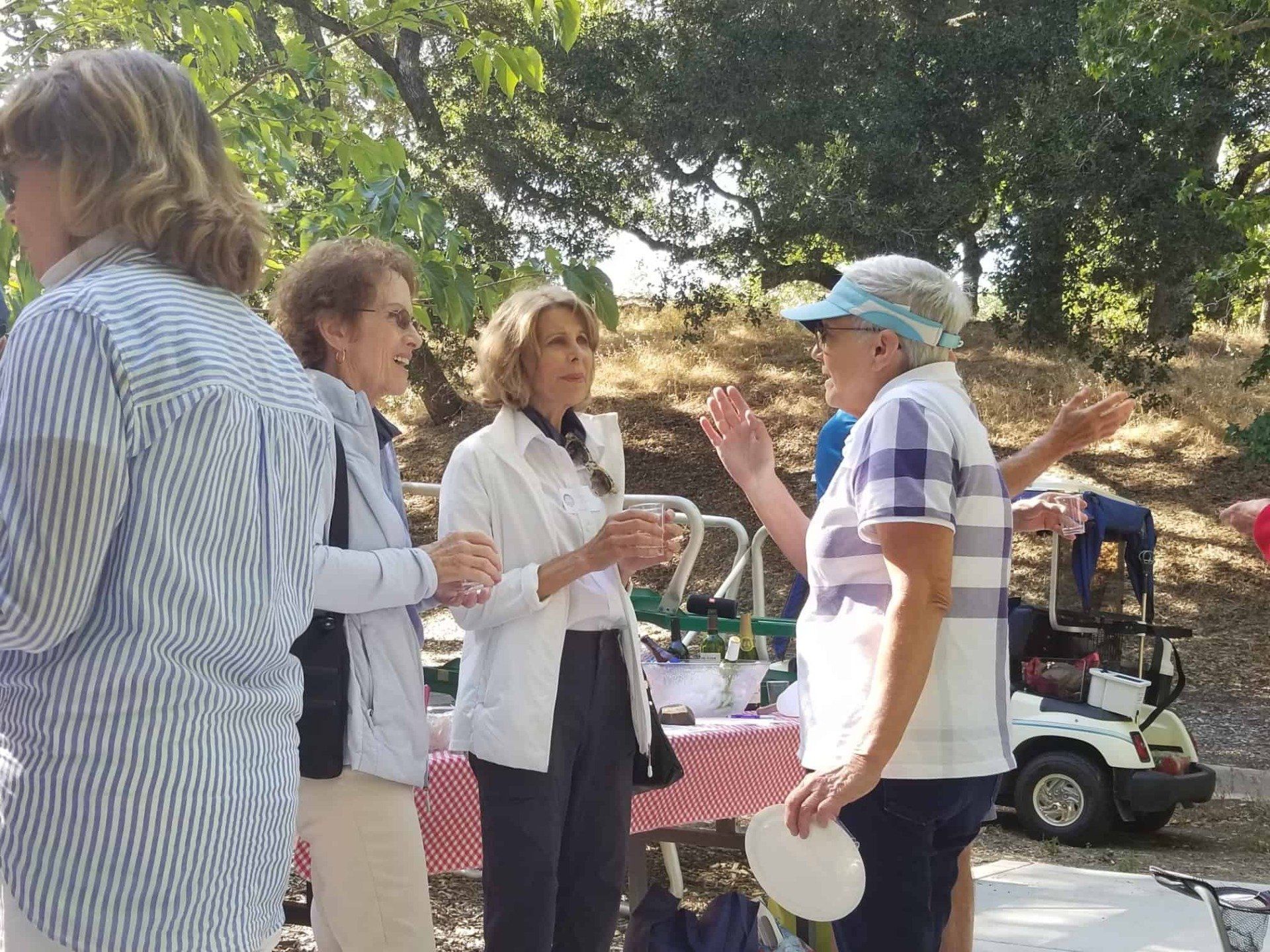 People chatting outdoors at a picnic table under trees, with a golf cart in the background.