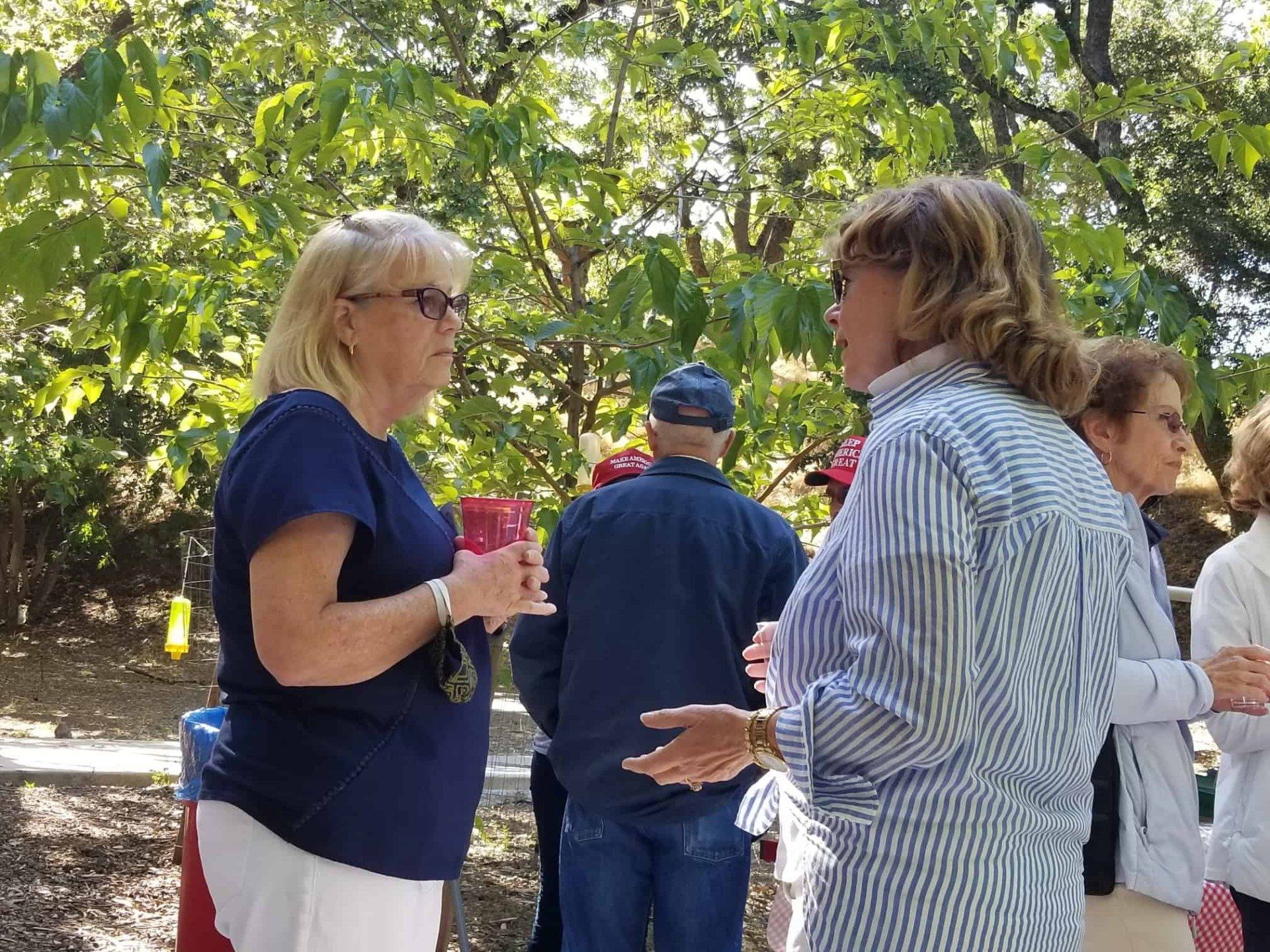 People chatting outdoors among trees, with a woman in a blue shirt holding a pink cup.