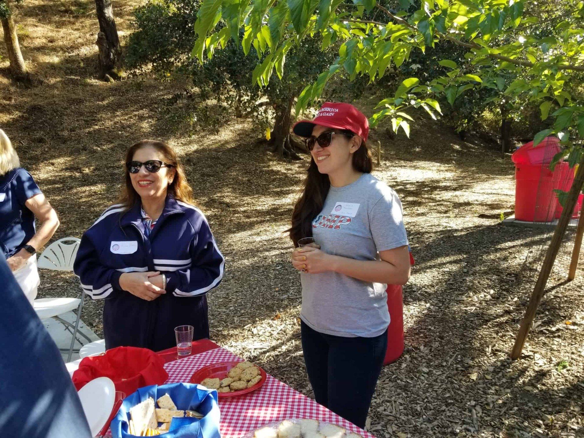 Two women stand at a picnic table outdoors, with plates and snacks under trees.