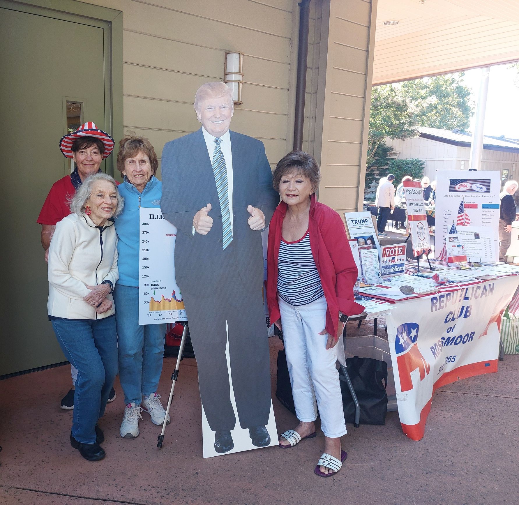 Group posing beside a life-size cardboard cutout at an outdoor community booth table with flyers and decorations