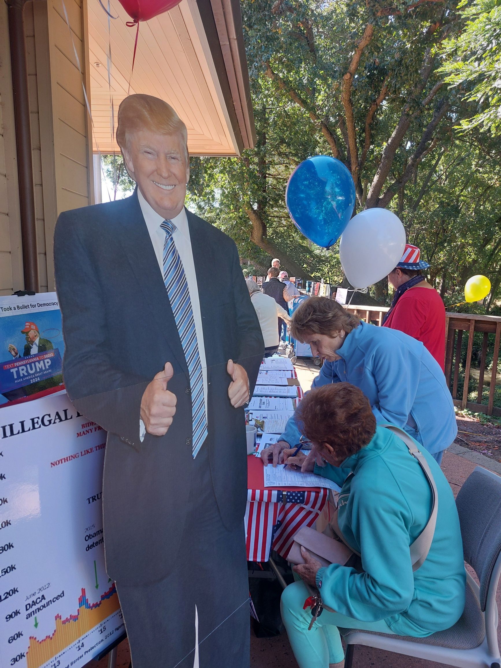 Cardboard cutout of Donald Trump with people at a table under blue and white balloons outdoors