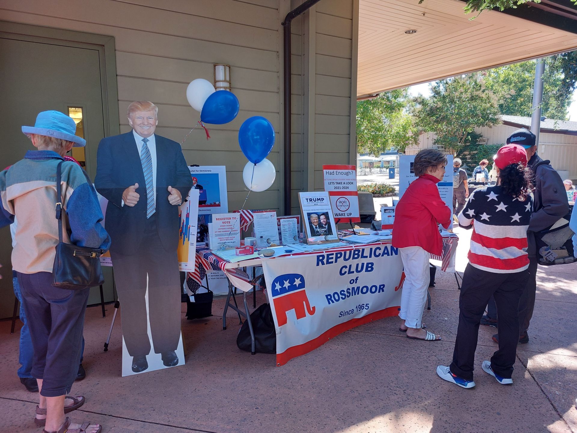 Political campaign table with cardboard cutout, blue balloons, and people talking on a porch