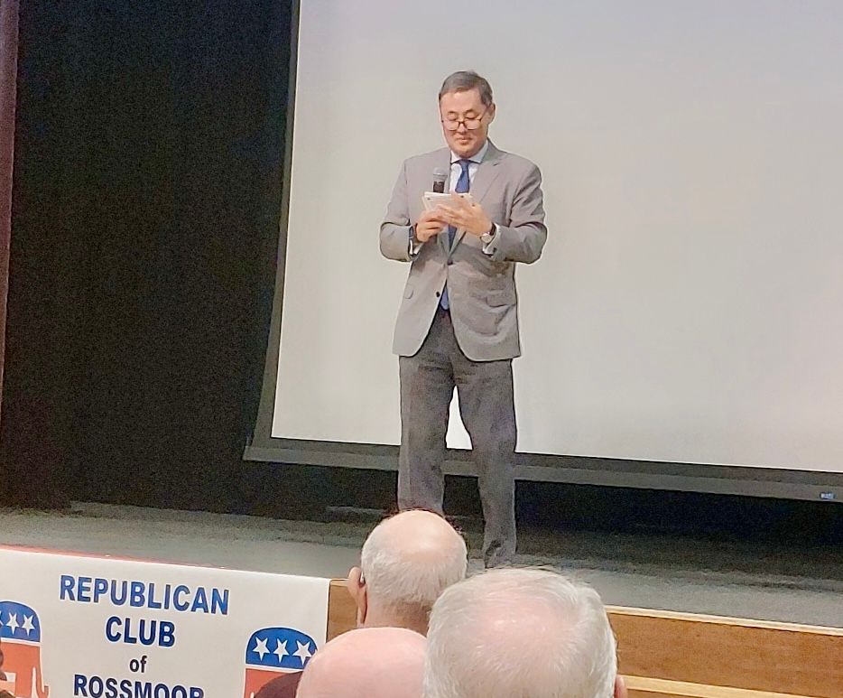 Man speaking on a stage before a projected screen, holding papers, with a Republican Club banner in front.