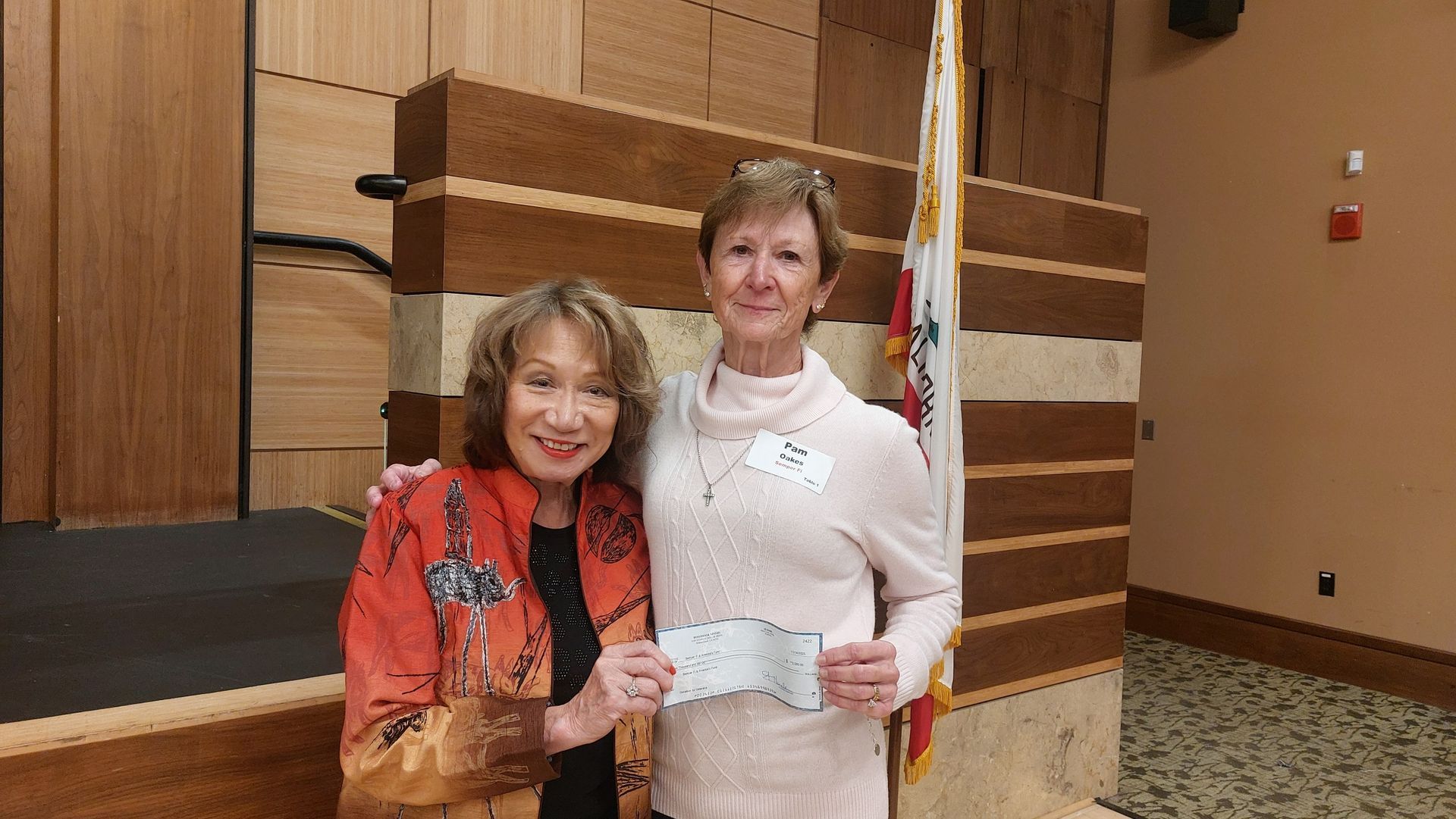 Two women smiling and holding a certificate in an auditorium, with an American flag in the background.