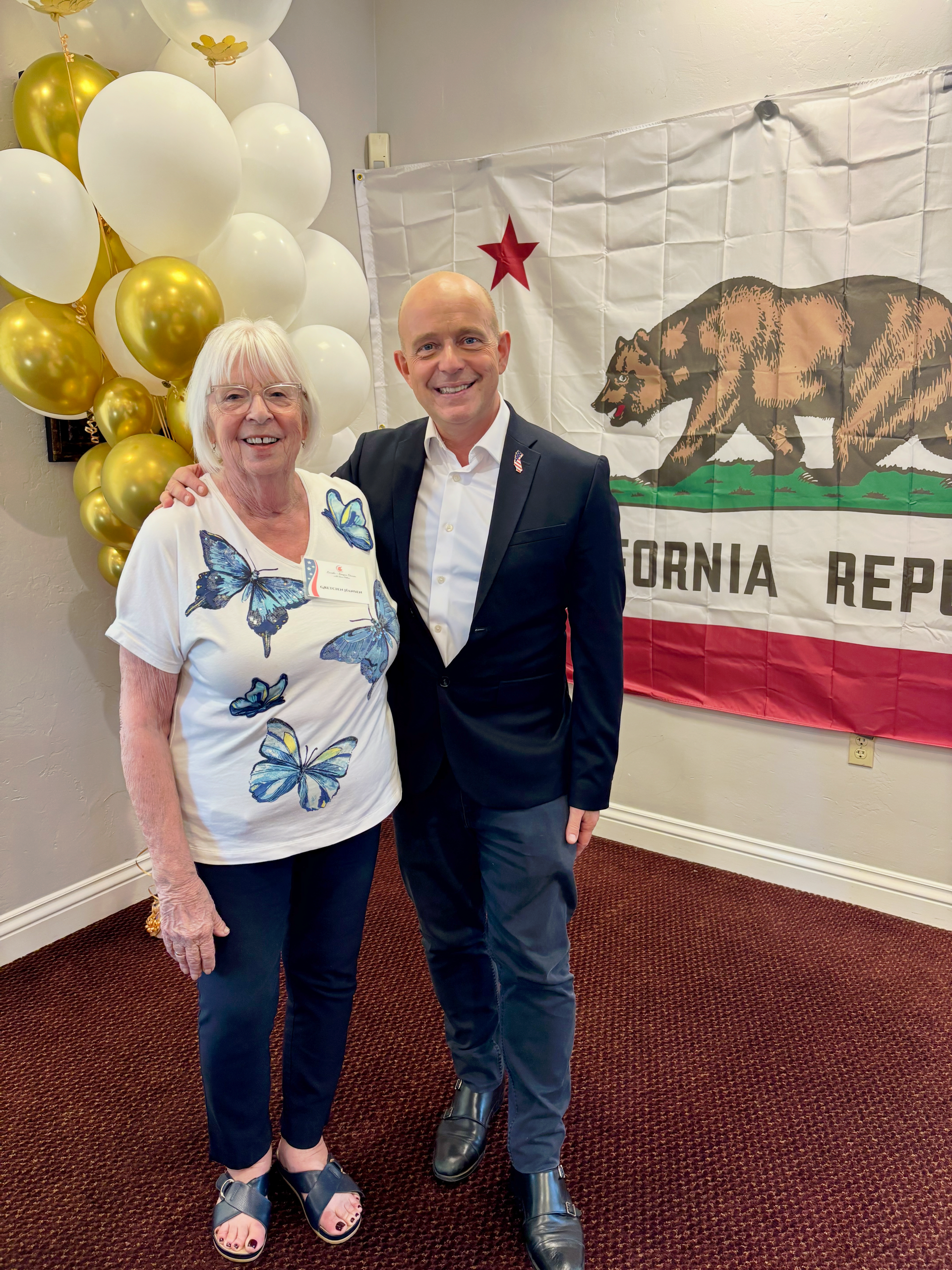 Two people pose indoors beside gold-and-white balloons and a California Republic flag backdrop.