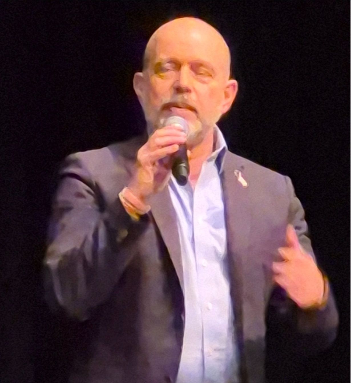 Man in a dark suit speaking into a microphone on stage under spotlight