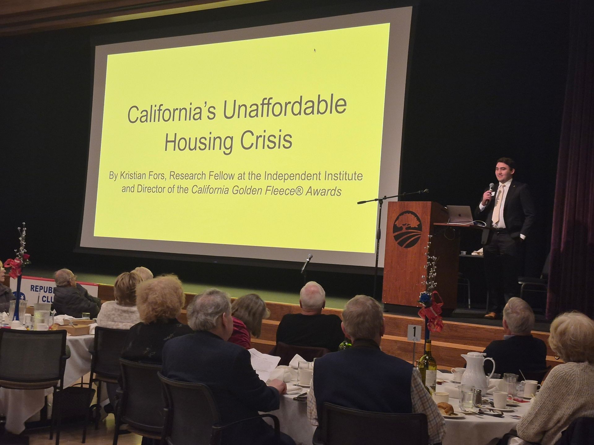 Conference presentation on California’s unaffordable housing crisis, with speaker at podium and audience seated at tables