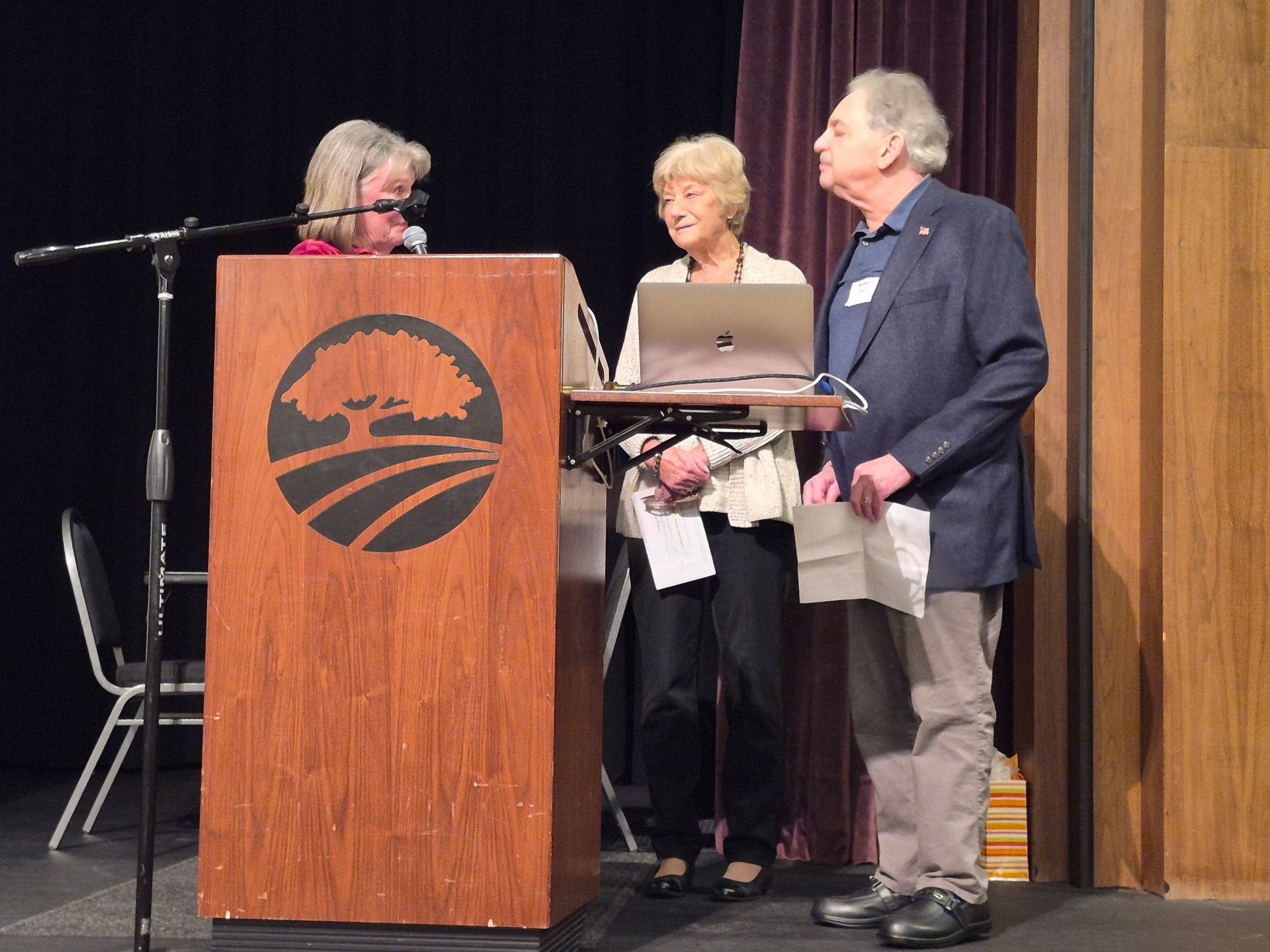Three people stand on stage beside a podium with a logo, one speaking into a microphone.