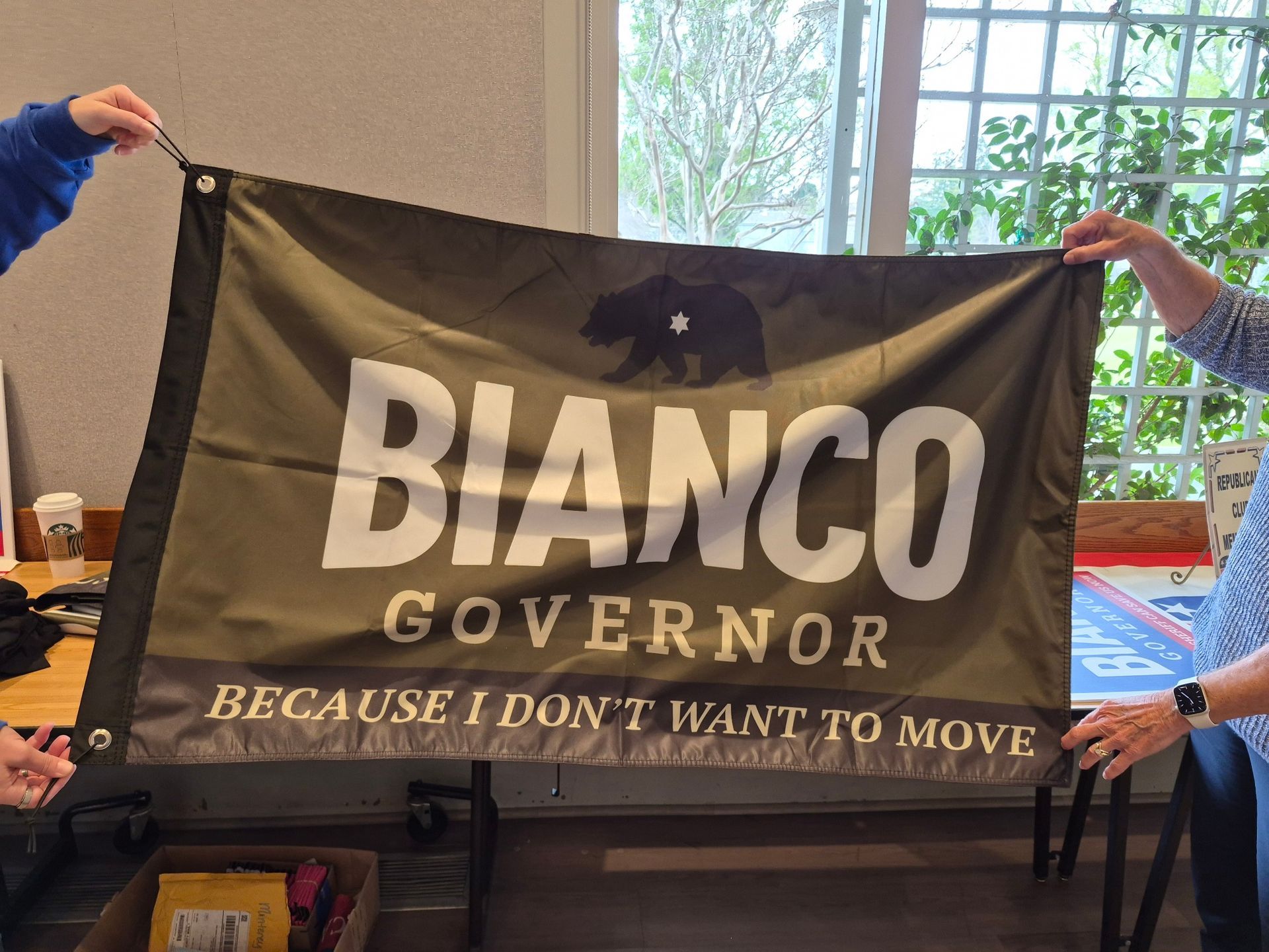 Black Bianco governor campaign banner held indoors by two people, with slogan “Because I don’t want to move.”