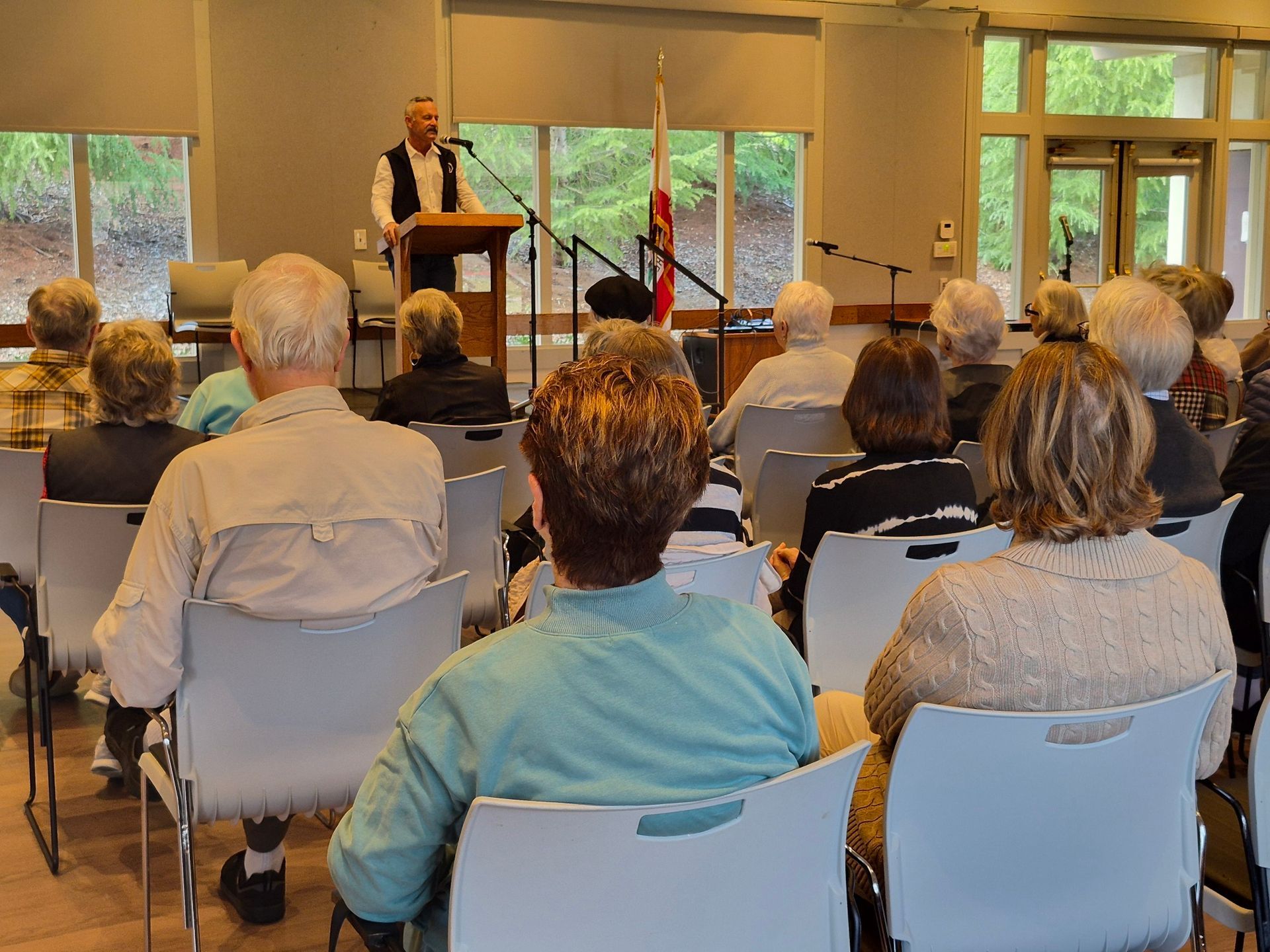 Audience seated in a hall listening to a speaker at a podium near large windows.